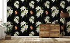 A room interior featuring a black wallpaper with a repeating pattern of large white lilies, green leaves, and small peach-orange birds. A light wood chest of drawers stands against the wall, with a floor lamp, rug, and potted plant in a bright room.