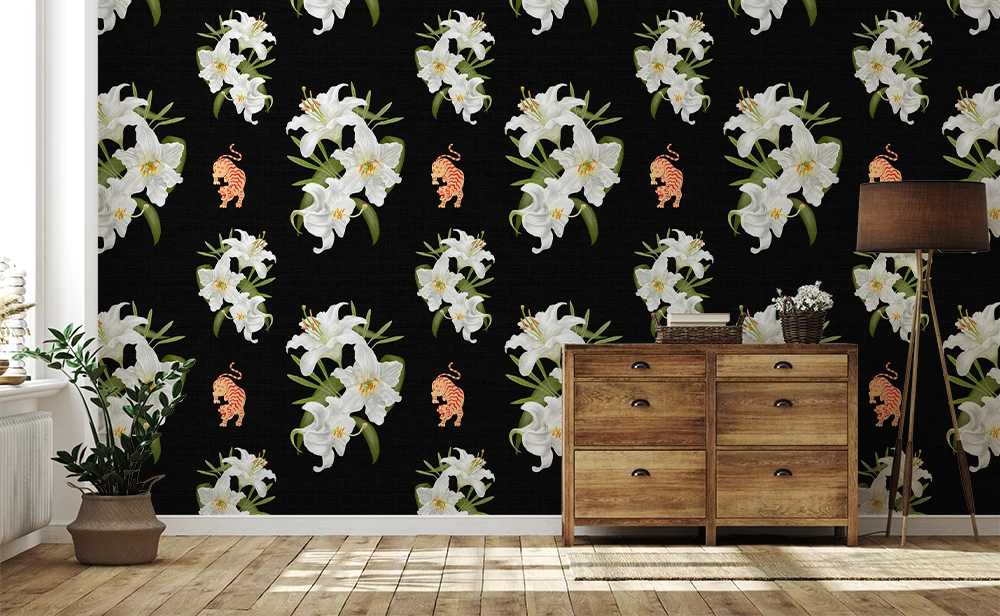 A room interior featuring a black wallpaper with a repeating pattern of large white lilies, green leaves, and small peach-orange birds. A light wood chest of drawers stands against the wall, with a floor lamp, rug, and potted plant in a bright room.