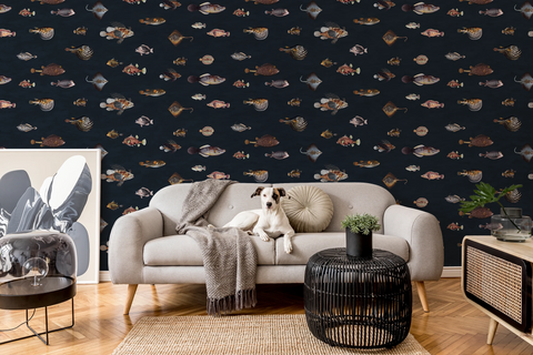 A styled living room featuring dark navy wallpaper with a repeating pattern of small illustrated fish in muted beige, brown, and gray tones. A light gray sofa with a dog lying on it, a black woven side table, and other room decor are visible in front of the wallpaper.