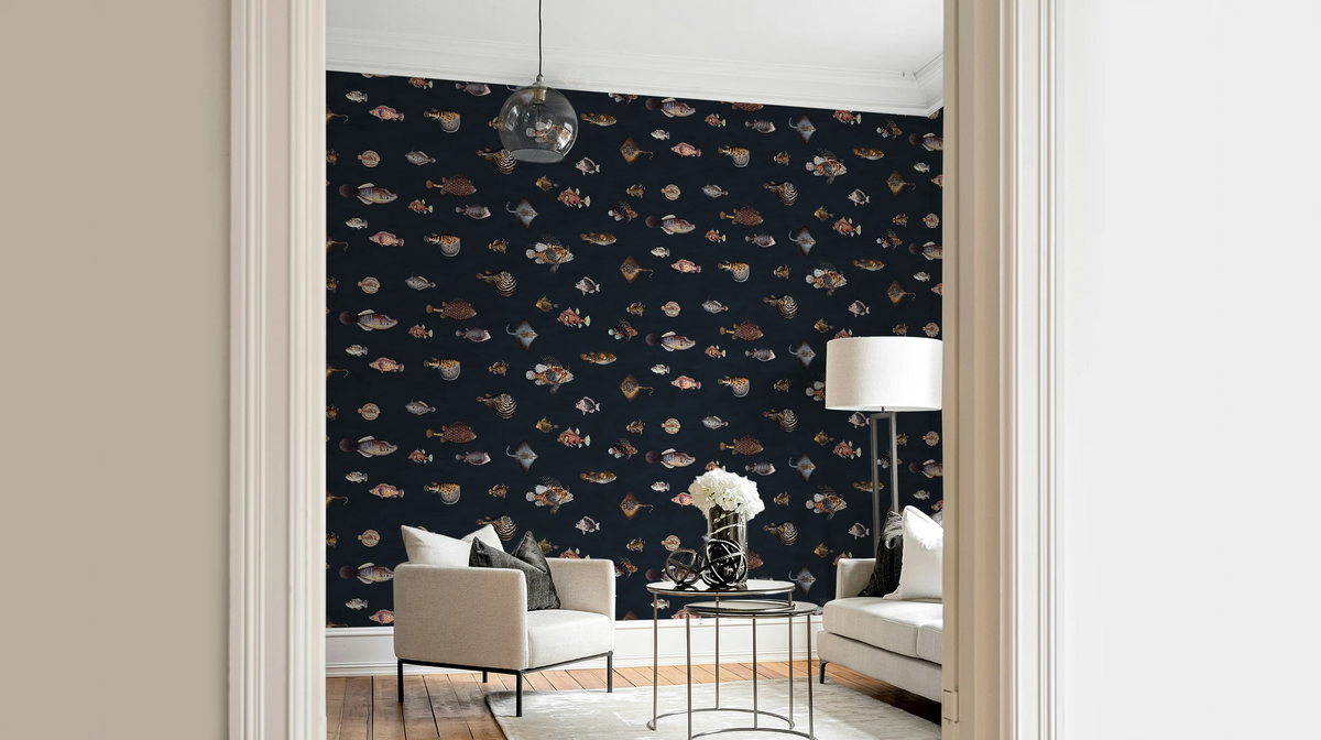 A styled interior room with a dark wall covered in a repeating fish pattern wallpaper, viewed through a doorway. The room contains a light beige armchair, a small round table with a vase of white flowers, a floor lamp, and a white sofa.