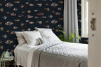 A styled bedroom featuring navy wallpaper with a pattern of vintage fish illustrations drawn by Marcus Elieser Bloch. It's a detailed pattern of various fish, including a large central fish and smaller striped, tropical, and mackerel fish. The bed is dressed in white and light gray bedding, with a window, gray curtains, and a green houseplant visible.