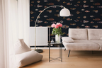 A styled living room featuring a dark navy wallpaper with a repeating pattern of colorful vintage fish illustrations drawn by Marcus Elieser Bloch. A beige sofa, a floor lamp, and a small side table with pink flowers.