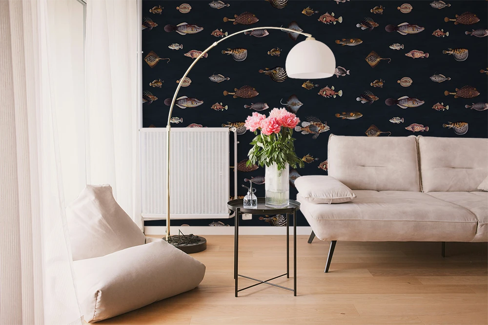 A styled living room featuring a dark navy wallpaper with a repeating pattern of colorful vintage fish illustrations drawn by Marcus Elieser Bloch. A beige sofa, a floor lamp, and a small side table with pink flowers.