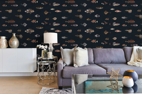 A styled living room featuring a dark navy blue wallpaper with a repeating pattern of various vintage fish illustrations drawn by Marcus Elieser Bloch, in muted beige, brown, and gray tones. In front of the wall is a gray fabric sofa with neutral throw pillows, a white sideboard with decorative vases, a small round metal side table with a lamp, and a glass coffee table in the foreground.