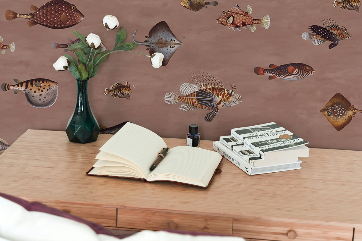 A dusty rose wallpaper with a pattern of vintage fish illustrations drawn by Marcus Elieser Bloch, a desk with books and a vase with white flowers. 