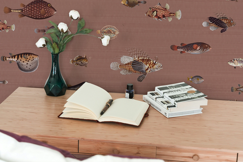 Small desk with a notepad, books, a vase with white flowers, and a wallpaper with a brown background featuring a pattern of vintage marine life illustrations. 