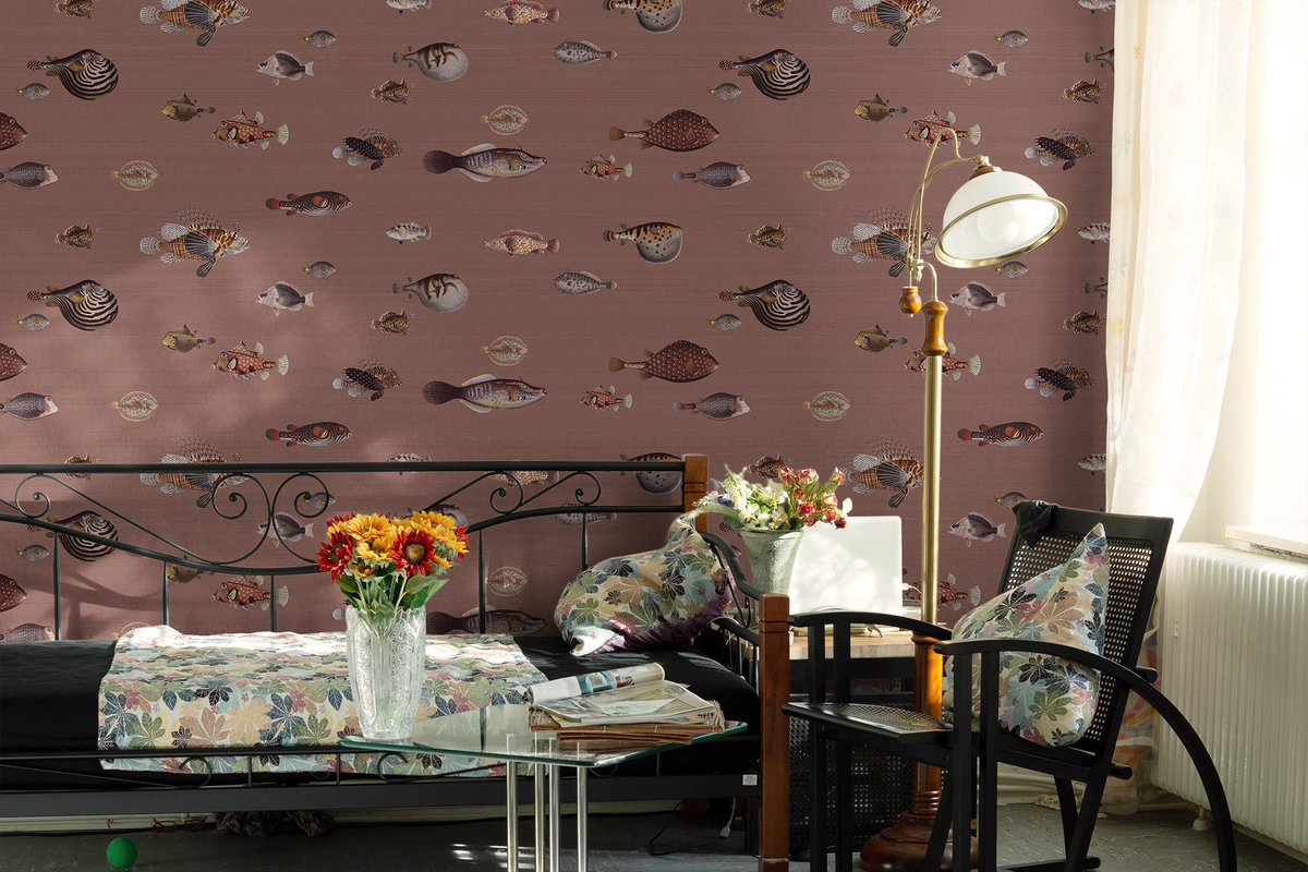 A styled bedroom interior featuring a dusty rose wallpaper with a repeating pattern of various illustrated fish. The room is furnished with a black metal daybed, a black armchair, a tall floor lamp, and floral accents.