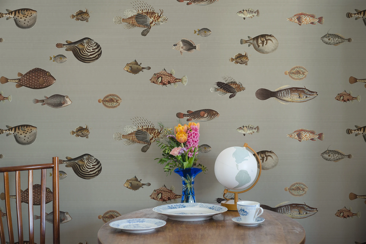 A small kitchen table in wood, with a chair, white plates, and a vase. The wall has a wallpaper with a greige background featuring a pattern of vintage marine life illustrations. 
