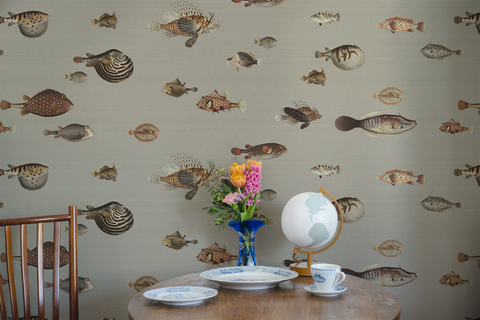 A small kitchen table in wood, with a chair, white plates, and a vase. The wall has a wallpaper with a greige background featuring a pattern of vintage marine life illustrations. 
