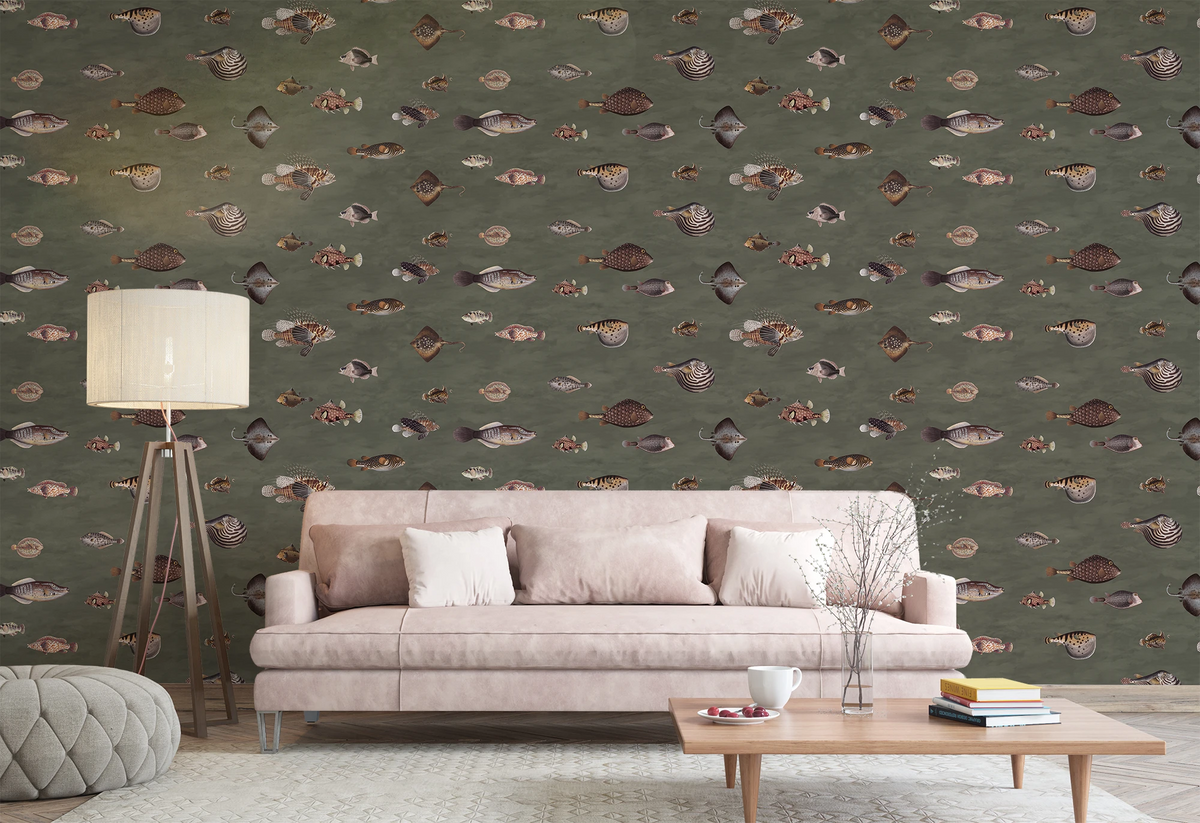 A styled living room featuring a sage green wallpaper with a repeating pattern of small fish in muted earthy tones. In front of the wall is a light pink sofa with matching pillows, a floor lamp, and a wooden coffee table.