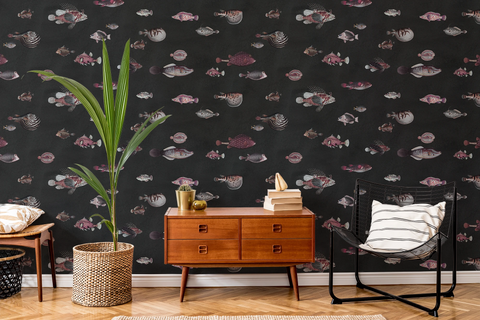 Mid century lounge living room, with a wooden side table, books, a black chair, and a plant with a textile pot. The wall features a wallpaper with a black background featuring a pattern of vintage marine life illustrations. 