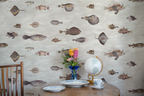 Fish-patterned wallpaper covering a wall in a styled room scene, with a wooden chair edge, round table, blue vase of flowers, globe, plates, and a teacup visible in the foreground.