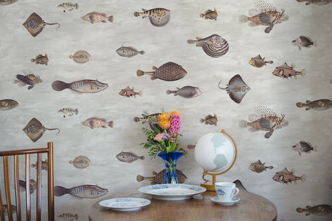 Fish-patterned wallpaper covering a wall in a styled room scene, with a wooden chair edge, round table, blue vase of flowers, globe, plates, and a teacup visible in the foreground.