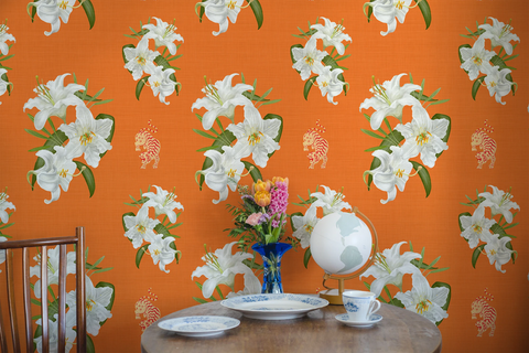 Bright Hermes orange wallpaper with a repeating pattern of large white lilies, green leaves, and small tiger motifs, shown in a styled interior scene with a round table, vase of flowers, globe, and teacup.