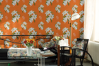 Bright bedroom scene featuring a Hermes orange wallpaper with large white floral prints, styled with a bed, chair, lamp, and floral accents.