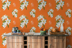 Bright kitchen scene with a Hermes orange wallpaper featuring large white lily floral patterns. A wooden counter with a dark stone top holds a coffee maker, mug, bowl of fruit, books, and a glass bottle.