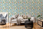 A styled living room featuring light Tiffany blue wallpaper with a repeating floral pattern with white lilies and orange tigers, a light gray sofa with a dog lying on it, a black woven side table, and a large framed abstract print.