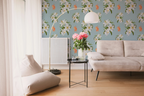 A styled living room corner featuring a light beige sofa, a matching floor cushion, a black side table with pink flowers, a gold floor lamp, and a light Tiffany blue wallpaper with a white floral pattern and orange tigers.