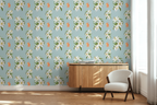A styled interior scene showing a wall covered in repeating floral wallpaper with white flowers, green leaves, and orange tigers, on a Tiffany blue background. A light wood sideboard and a white upholstered chair sit in front of the wall, with a window and curtains to the right.