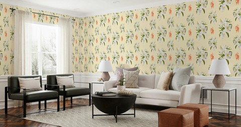 A styled living room featuring floral wallpaper with a cream background, orange tigers, and green leaves. The room includes a cream sofa with decorative pillows, two black leather armchairs, a round black coffee table, side tables with lamps, and brown leather ottomans.