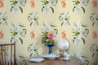 Small wooden table with porcelain tea cups, white plates, flowers, a globe, and a wallpaper with a pattern of white lilies and orange tigers. 
