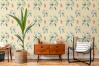 A styled interior room featuring wallpaper with a repeating pattern of white flowers, green leaves, and small orange tigers on a pale yellow background. A mid-century wooden sideboard, a black wire chair with a striped pillow, a tall potted plant, and a small table are arranged in front of the wall.