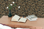 A wooden desk, with books, a notebook and pen, a vase, and the wall behind it  features a tapestry wallpaper with an oak leaf pattern. 