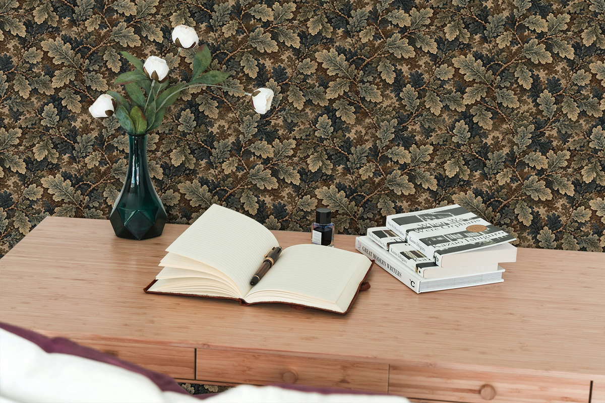 A wooden desk, with books, a notebook and pen, a vase, and the wall behind it  features a tapestry wallpaper with an oak leaf pattern. 
