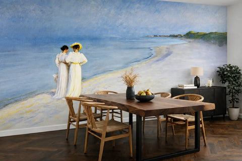 A large art wallpaper mural depicting a beach scene with two women in white dresses stands on the wall behind a wooden dining table and chairs in a modern dining room. The painting is "Summer Evening on the Southern Beach" by Peder Severin Krøyer.