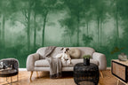 A dog sits on a light beige sofa in a styled living room with a large green forest mural wallpaper on the wall behind it. The room includes a black woven side table, a small side table with a plant, and a wooden floor.
