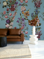 A styled interior scene featuring a large floral wallpaper mural in muted blue, pink, purple, and green tones behind a brown leather sofa, with a white vase and dried flowers on a white pedestal in the foreground.