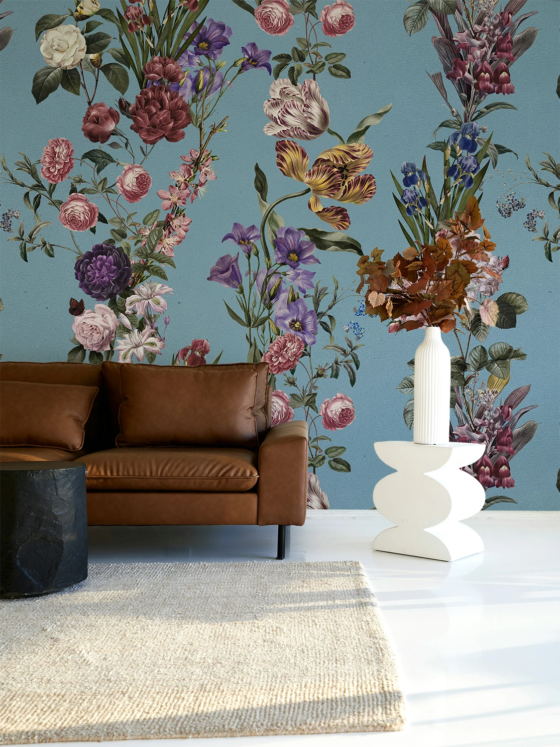 A styled interior scene featuring a large floral wallpaper mural in muted blue, pink, purple, and green tones behind a brown leather sofa, with a white vase and dried flowers on a white pedestal in the foreground.