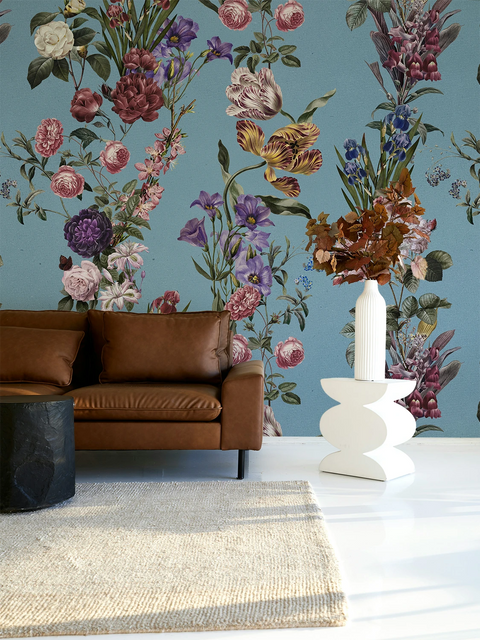 A styled interior scene featuring a large floral wallpaper mural in muted blue, pink, purple, and green tones behind a brown leather sofa, with a white vase and dried flowers on a white pedestal in the foreground.