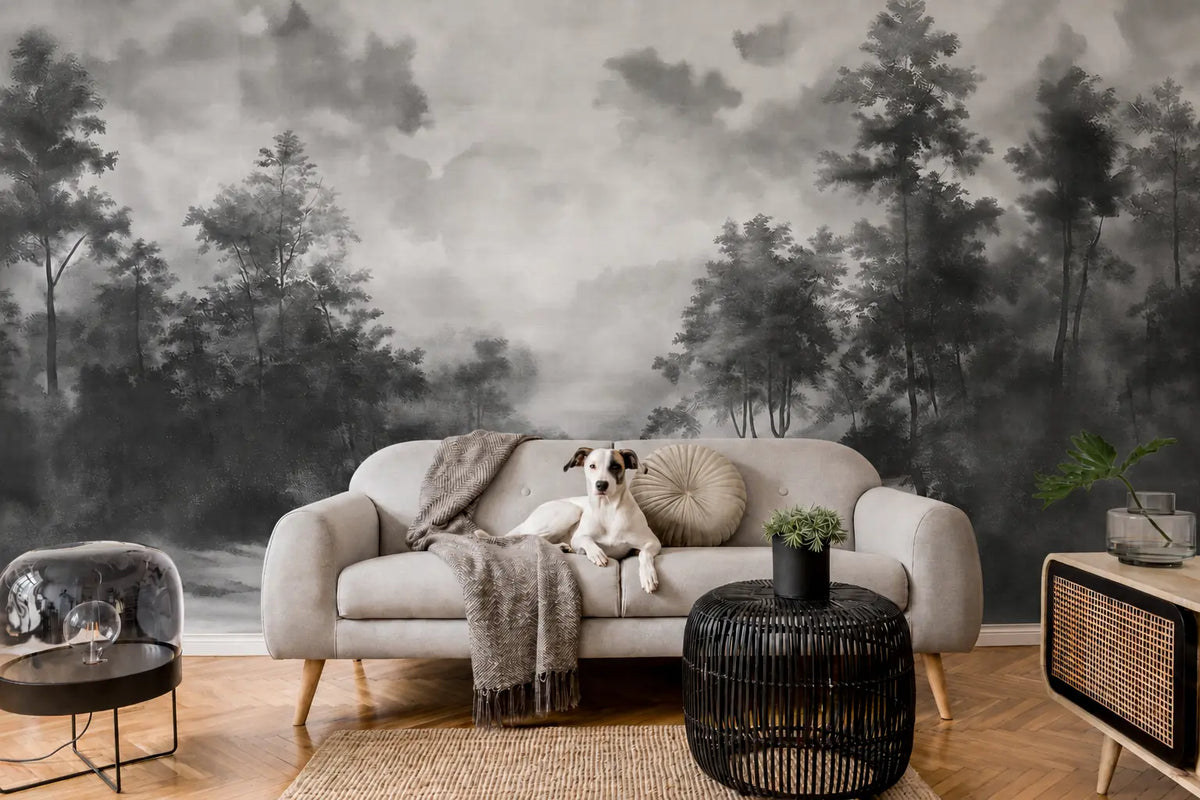 A modern living room featuring a large-scale black and white misty forest wallpaper mural. A light gray fabric sofa sits in the center with a small white and black dog lying on it, draped with a gray throw blanket.