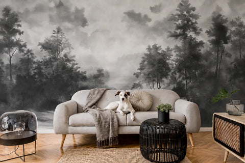 A modern living room featuring a large-scale black and white misty forest wallpaper mural. A light gray fabric sofa sits in the center with a small white and black dog lying on it, draped with a gray throw blanket.