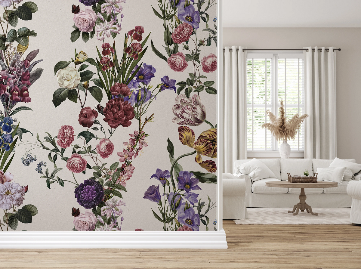 A wall covered in a dense botanical floral wallpaper pattern with pink, purple, white, and green flowers on a light beige background, shown in a styled living room next to a white sofa and window.
